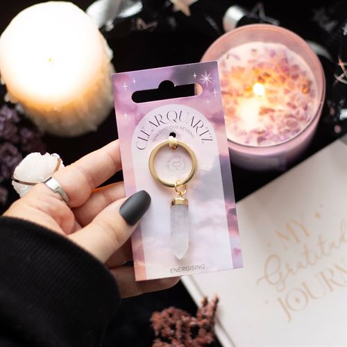 A hand holding a Clear Quartz Crystal Keyring displayed on celestial-themed packaging, surrounded by candles and a gratitude journal. This high-vibration crystal amplifies energy, enhances spiritual awareness, and serves as a personal talisman for manifestation and protection.