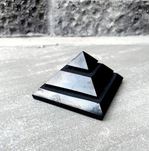 A Sakkara Shungite Pyramid with a stepped design, placed on a stone surface, known for its EMF protection and grounding energy.