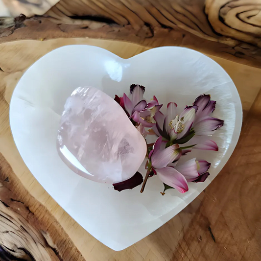 Selenite heart-shaped bowl with rose quartz crystal and fresh flowers, placed on a wooden surface. A sacred altar dish for crystal cleansing, rituals, and spiritual offerings.